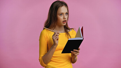 pensive teenage girl holding notebook and pen isolated on pink