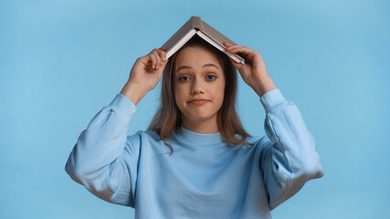 teenage girl in soft sweatshirt holding book above head isolated on blue