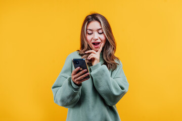 Surprised happy girl looks at the phone, she is wearing a sweater, behind her a yellow background with space for text