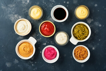 Set of sauces in bowls - ketchup, mayonnaise, mustard, soy sauce, bbq sauce, pesto, chimichurri, mustard grains on dark stone background. Top view copy space.