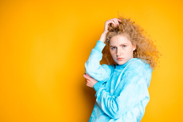 A young girl with her hand straightens her blond hair while standing on a bright background