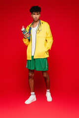 full length of curly african american man in yellow rain jacket standing with skipping rope and sports bottle on red