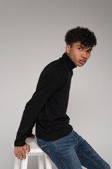 african american young man in turtleneck sweater leaning on white chair isolated on grey