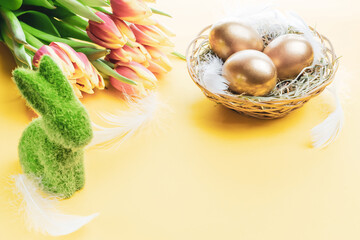 Golden easter colour eggs in basket with spring tulips, white feathers on pastel yellow background in Happy Easter decoration. Spring holiday concept.