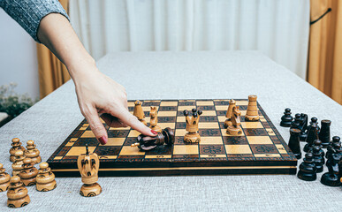 Un tablero de ajedrez, de madera, con fichas blancas y una mano de mujer que tumba al rey negro