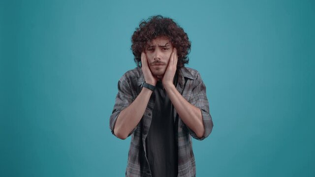 The young hipster, puzzled, with his bread on his face, looks into the room and can't believe it. Studio image on blue background. Young man dressed in a shirt and a T-shirt. Curly-haired teenager. Hi