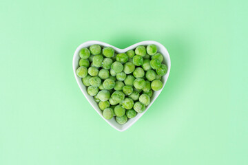 Green frozen peas in a white heart-shaped bowl on light green background. Concept love vegetables,...