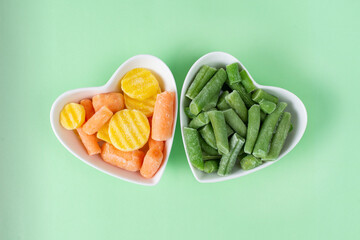 Green frozen asparagus beans, baby carrot mix in white heart-shaped bowl, light green background. Concept love vegetables, vegetarian food. Diet ingredient. Healthy meal prep to freezer blank