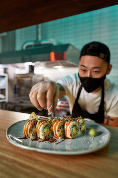 Professional sushi chef wearing protective mask and gloves decorating sushi rolls preparing to be served on plate at commercial kitchen