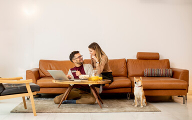 Young woman and young man using laptop for onlin payment while sitting by sofa with their shiba inu...