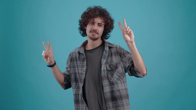 Young hipster with the fingers of both hands shows the sign of peace. Smile wide. Young man dressed in a shirt and a T-shirt. Curly-haired teenager. Hipster carizmaticv