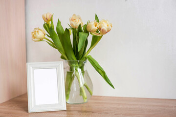 Bouquet of beautiful tulips and empty frame mock up. Copy space. International Women's Day celebration. White wall background. Scandinavian interior.