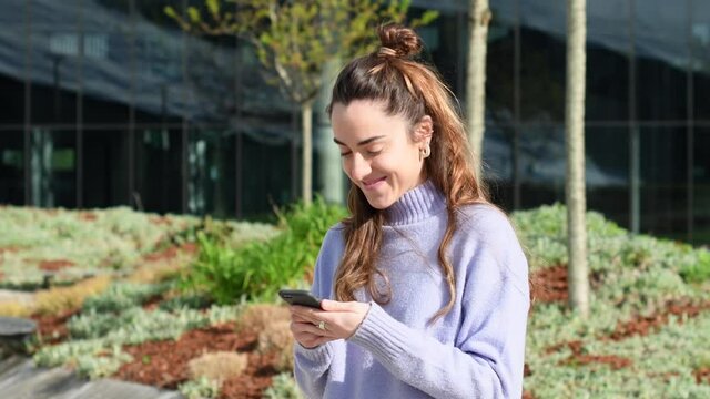 Static scene of a pretty young woman receiving good news on her cell phone and smiling outdoors in an urban park.