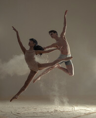 Two ballet dancers perform splits and twine poses in jump above the floor covered flour.