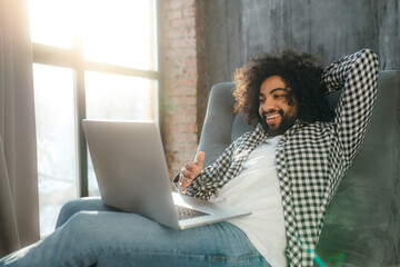 Smiling Egyptian in a plaid shirt sits in a chair working at the computer. Work remotely