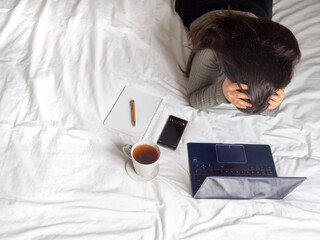 Frustrated woman working from home lying on bed with white sheets. Stressed female studying online with coffee in bed and a laptop. Top view of defeated girl working remotely during pandemic.