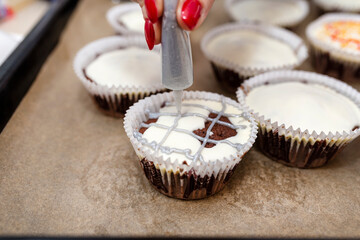 A woman squeezes colored frosting from a tube onto chocolate brown cupcakes covered with white frosting with colorful decorations.