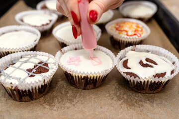 A woman squeezes colored frosting from a tube onto chocolate brown cupcakes covered with white frosting with colorful decorations.
