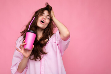 Shot of beautiful sexy young positive brunette curly woman wearing pink shirt isolated over pink background holding paper coffee cup for mockup drinking and enjoying