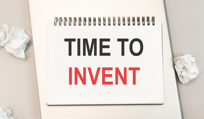 The word time to invent is written in a notebook that sits on a gray desktop along with a laptop....
