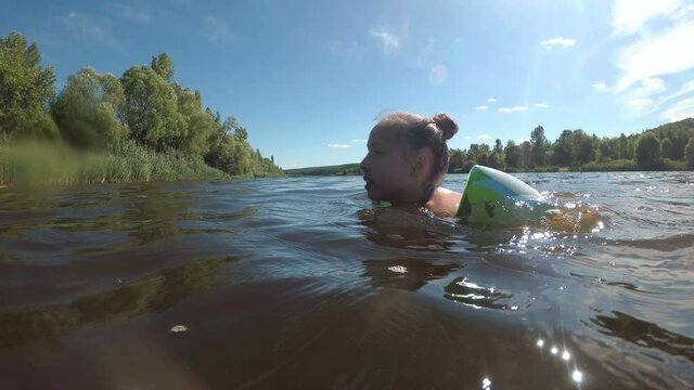 A child, a little girl, learns to swim in the river and swims through the water in inflatable armbands on a sunny summer day. The concept of a healthy lifestyle. Summer holidays. Close-up. 4K