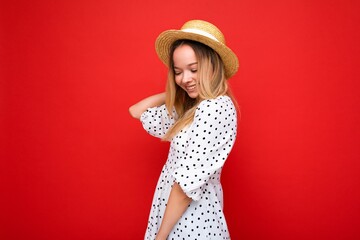 Portrait of young beautiful smiling hipster blonde woman in trendy summer dress and straw hat. Sexy carefree female person posing isolated near red wall in studio. Positive model with natural makeup