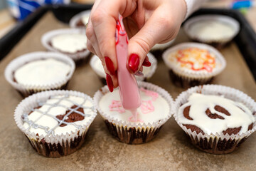 A woman squeezes colored frosting from a tube onto chocolate brown cupcakes covered with white frosting with colorful decorations.