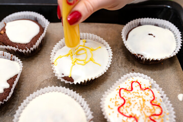 A woman squeezes colored frosting from a tube onto chocolate brown cupcakes covered with white frosting with colorful decorations.