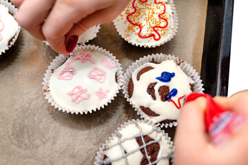Fototapeta premium A woman squeezes colored frosting from a tube onto chocolate brown cupcakes covered with white frosting with colorful decorations.