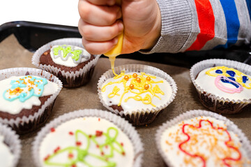 A child squeezes colored frosting from a tube onto chocolate brown cupcakes covered with white frosting with colorful decorations, isolated on white.