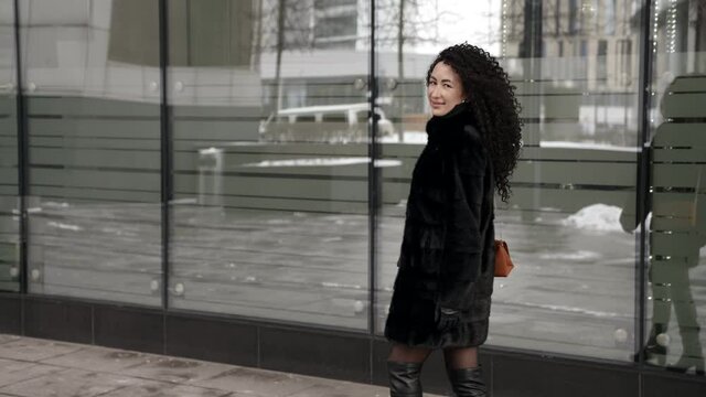 stunning pretty woman with long curly hair is walking alone on city street at winter day, dressed natural fur coat