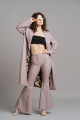 Studio shot of pretty fashion model in pastel purple leather trousers, coat, khaki sunglasses on gray background
