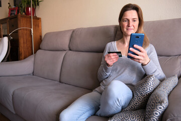 Happy woman doing shopping online sitting on couch at home. Lady insert credit card number on website with her smart phone.