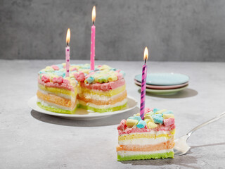 Rainbow cake decorated with festive candles on a gray background. A cut - off piece of birthday cake. Festive mood. Three years old. Space for text. Copy space. Colorful children's dessert. Holiday.
