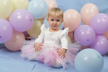 A beautiful little girl 1 year old in white turtleneck and pink tutu skirt sits among the balloons and looks at the camera