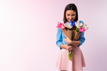 Studio shot of good looking woman smells tulip flowers, enjoys pleasant odor, stylish clothes indoor. Lovely caucasian lady hold beautiful bouquet of spring flowers. Neon pink background