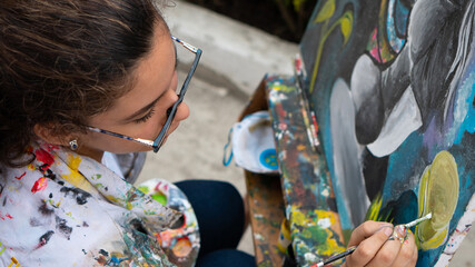 Hispanic young woman painting on an incomplete canvas, she wears glasses due to myopia. Curly brown...