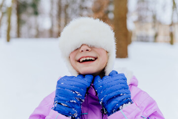 Winter portrait happy smiling young preteen girl having fun pulls a hat over her eyes wearing winterwear and white hat with fluffy fur on snowy park background.