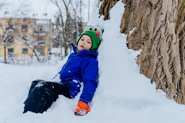 Winter baby. Five year old cute kid dressed in warm clothing sitting at snow lean on big tree. Tired child relaxing after play in cold frosty day, outdoor.