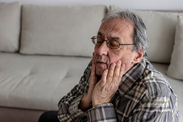 Elderly man having toothache, touching his cheek, suffering from pain while sitting on sofa at home. Depressed ill man having toothache and touching cheek. Mature man suffering from tooth pain, caries