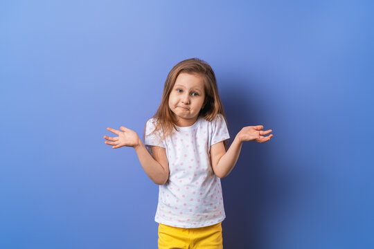 Small Sad Blonde Child A Guilty 5-year-old Girl In A T-shirt Spreads Her Hands Oops Gesture On A Purple Background In The Studio. The Concept Of Children's Lifestyle. Bad Behavior.