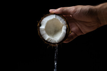 Fototapeta premium Isolated coconut on a black background. Coconut milk is poured from half a coconut. Half of a coconut in a man's hand on a dark background. Healthy fruit