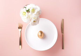 On a white plate lies a golden egg, next to white flowers, and golden cutlery on a powdery background, horizontal orientation, copy space