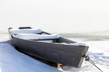 Snow-covered old wooden rowing boat on the shore in winter season. Fishing boat. A frozen lake or river. Low ambient temperature. The season of winter fishing. Sunny winter day.