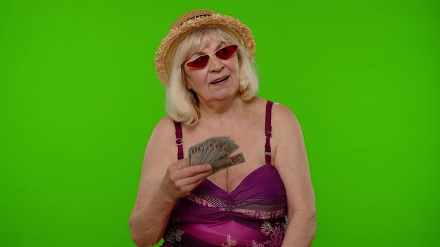 Rich senior woman tourist with cash fan enjoying financial independence, waving heap of currency dollar bills on chroma key background. Travel, summer holiday vacation. Elderly grandmother in swimsuit