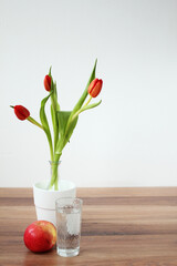 red tulips on a wooden table in a vase with an apple and a glass of water