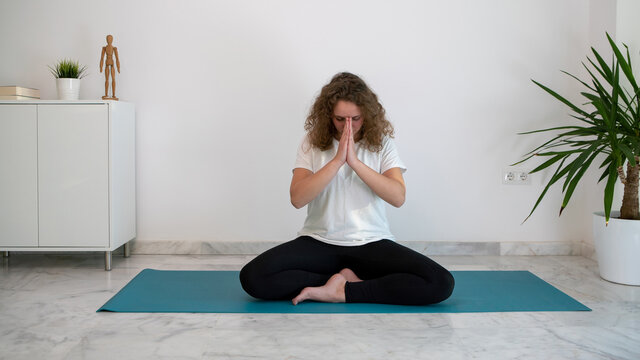 Young woman practicing yoga and doing the Namaste salute on a blue mat at home.