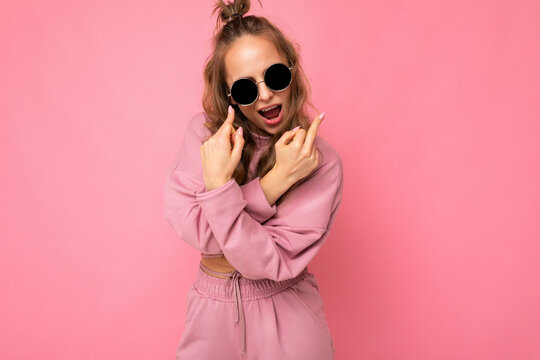 Young self-confident positive cool beautiful blonde wavy-haired woman with sincere emotions wearing stylish pink sport suit and sunglasses isolated on pink background with copy space and showing fuck