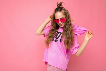Charming positive funny young blonde curly woman isolated on pink background wall wearing casual pink t-shirt and stylish colourful sunglasses looking at camera