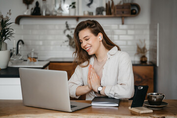 Young pretty woman talking on video call and waving hand while sitting at table inthe kitchen